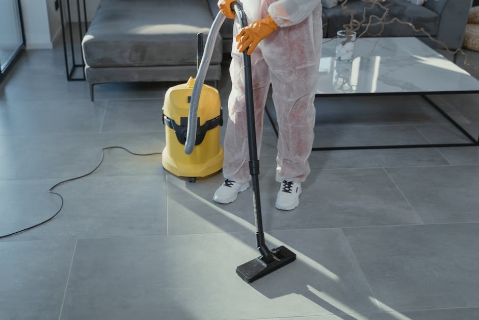 A person wearing a light green cleaning glove is reaching for a set of white binders or file folders on a dark wooden shelf, which is located in a room with beige walls. The shelf contains a few hardc