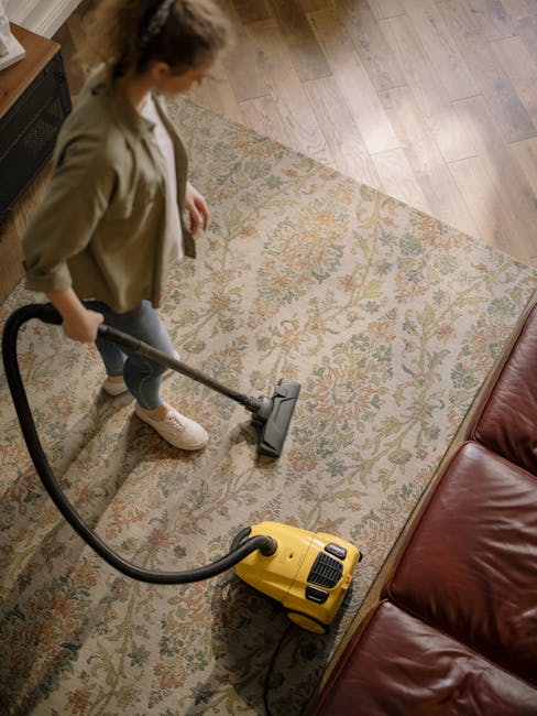 A woman using a yellow and black vacuum cleaner to deep clean a traditional patterned area rug with beige, green, and orange floral motifs, situated on a wooden floor in a living room. The scene is li
