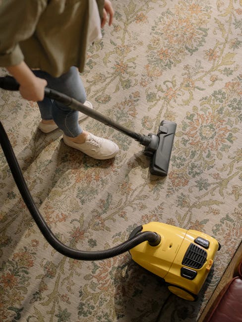 A woman using a yellow and black vacuum cleaner to deep clean a traditional patterned area rug with beige, green, and orange floral motifs, situated on a wooden floor in a living room. The scene is li