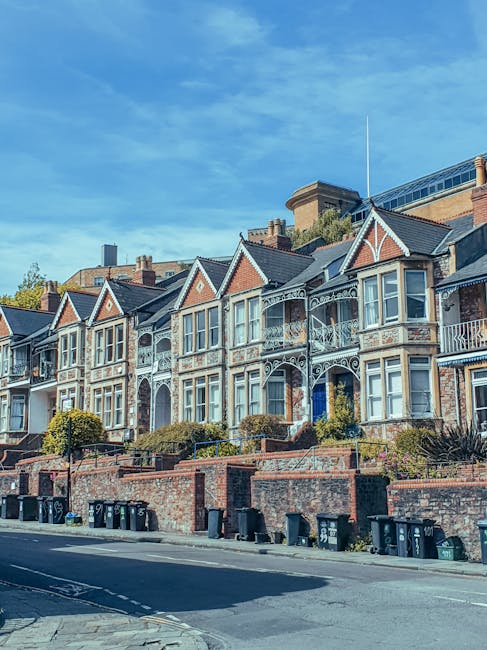 Photograph of a row of Victorian terrace houses with brick facades and white-framed sash windows, lining a paved sidewalk. The street appears quiet with some fallen leaves scattered on the pavement. T