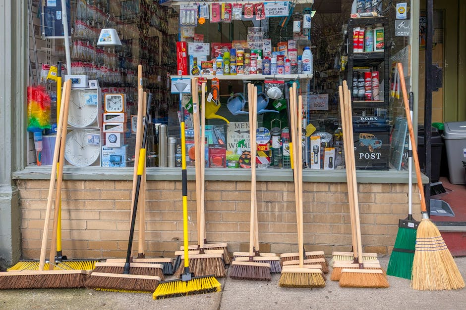 A row of various cleaning tools lined up in front of a store window, including brooms, mops, and dustpans with wooden handles, some with yellow bristles and others with natural brown or green syntheti